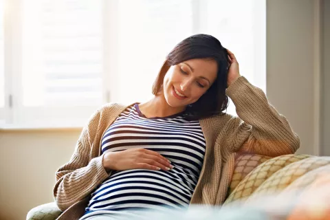 Pregnant adult with hand over stomach while sitting on the couch.