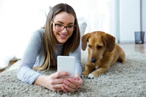 A young adult laying on the floor with a dog while looking a cell phone.