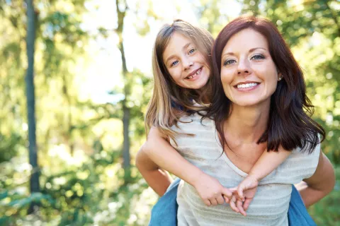 an adult giving a child a piggyback ride outside, both smiling at camera