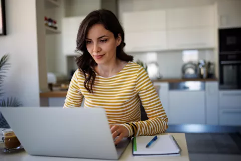 An adult typing on a laptop with a notebook on the side.