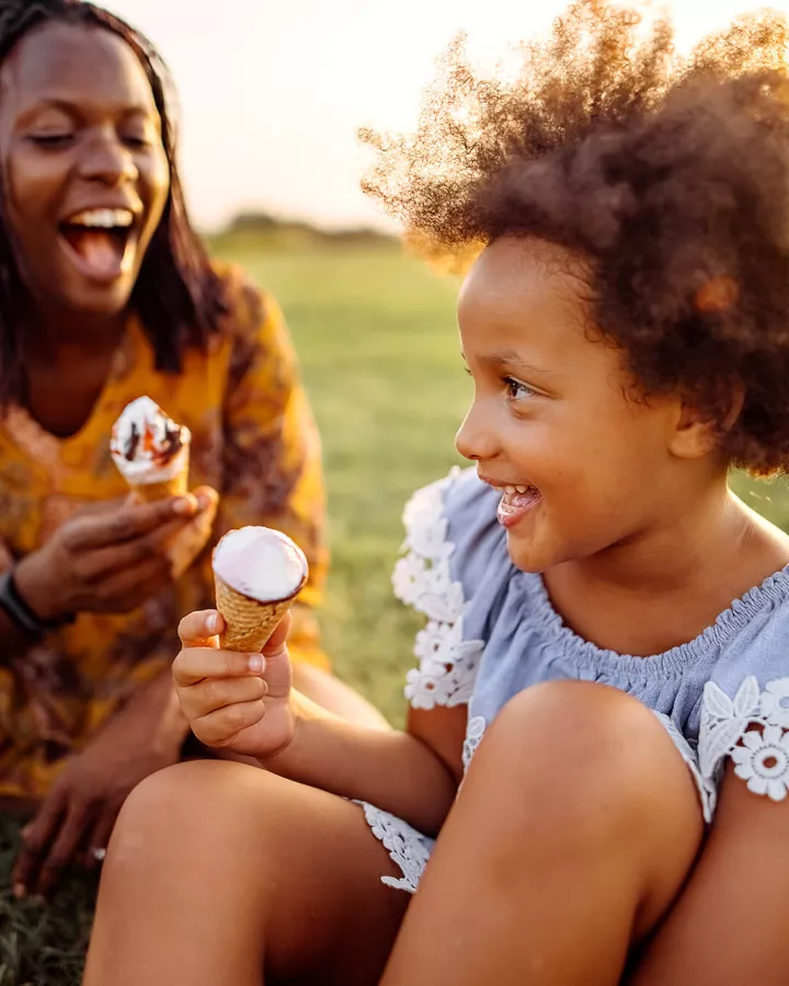 child and young adult eating ice cream in a field during sunset.