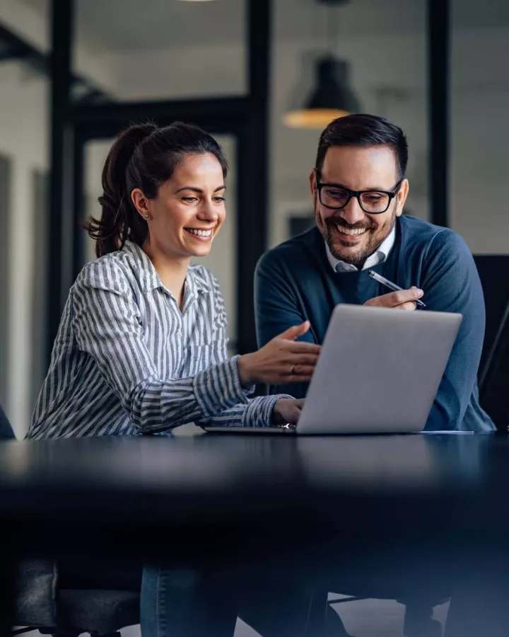 Two adults at a desk, smiling at a laptop.