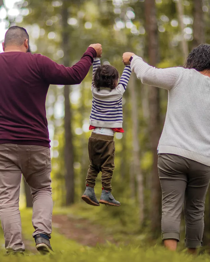 two adults lifting a child by the hands between them, hiking outdoors, facing away from camera