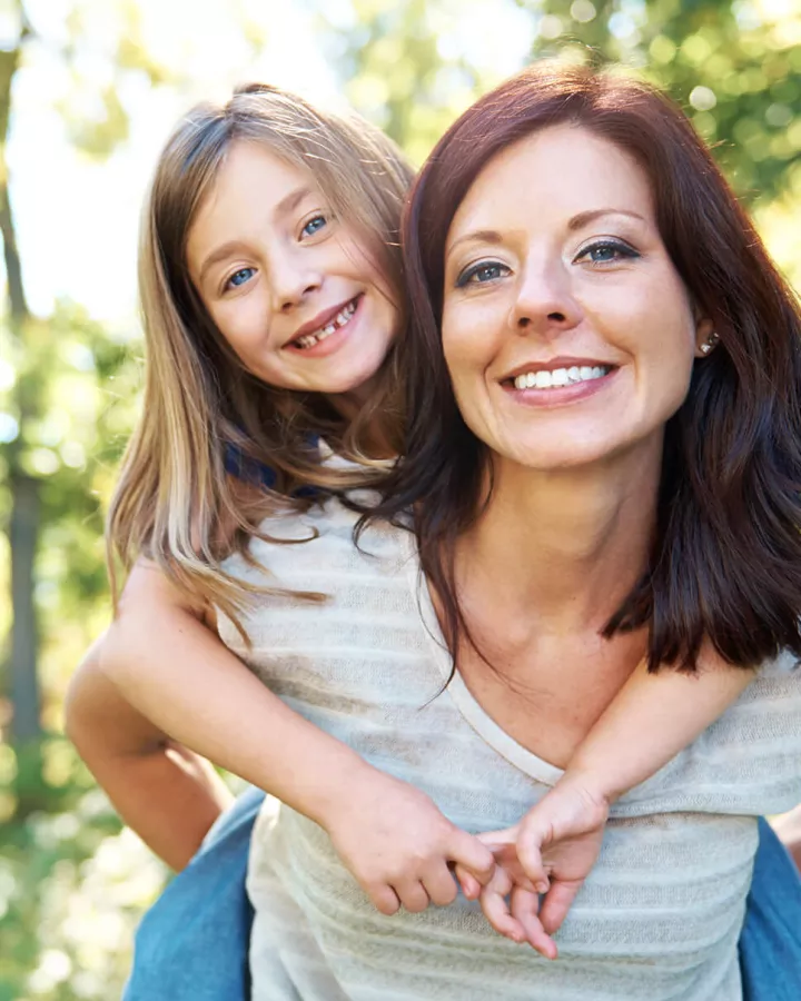 an adult giving a child a piggyback ride outside, both smiling at camera